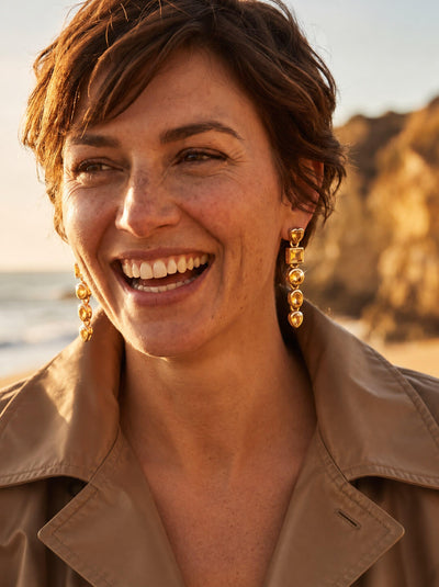 Citrine Symphony Earrings with golden drop settings and sparkling round citrine stones worn by a smiling woman on a sunlit beach