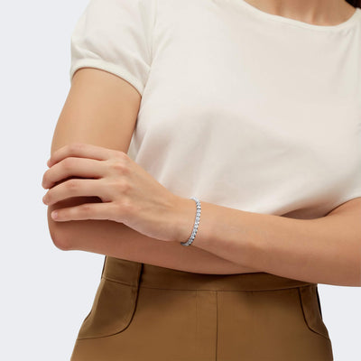 Woman wearing a Millionaire Rhodium Tennis Bracelet made of sterling silver with rhodium plating, styled elegantly on her wrist paired with a white top and brown pants.