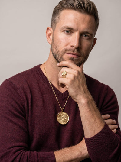 Man wearing a gold American Glory Ring and matching gold pendant necklace dressed in a burgundy sweater against a neutral background