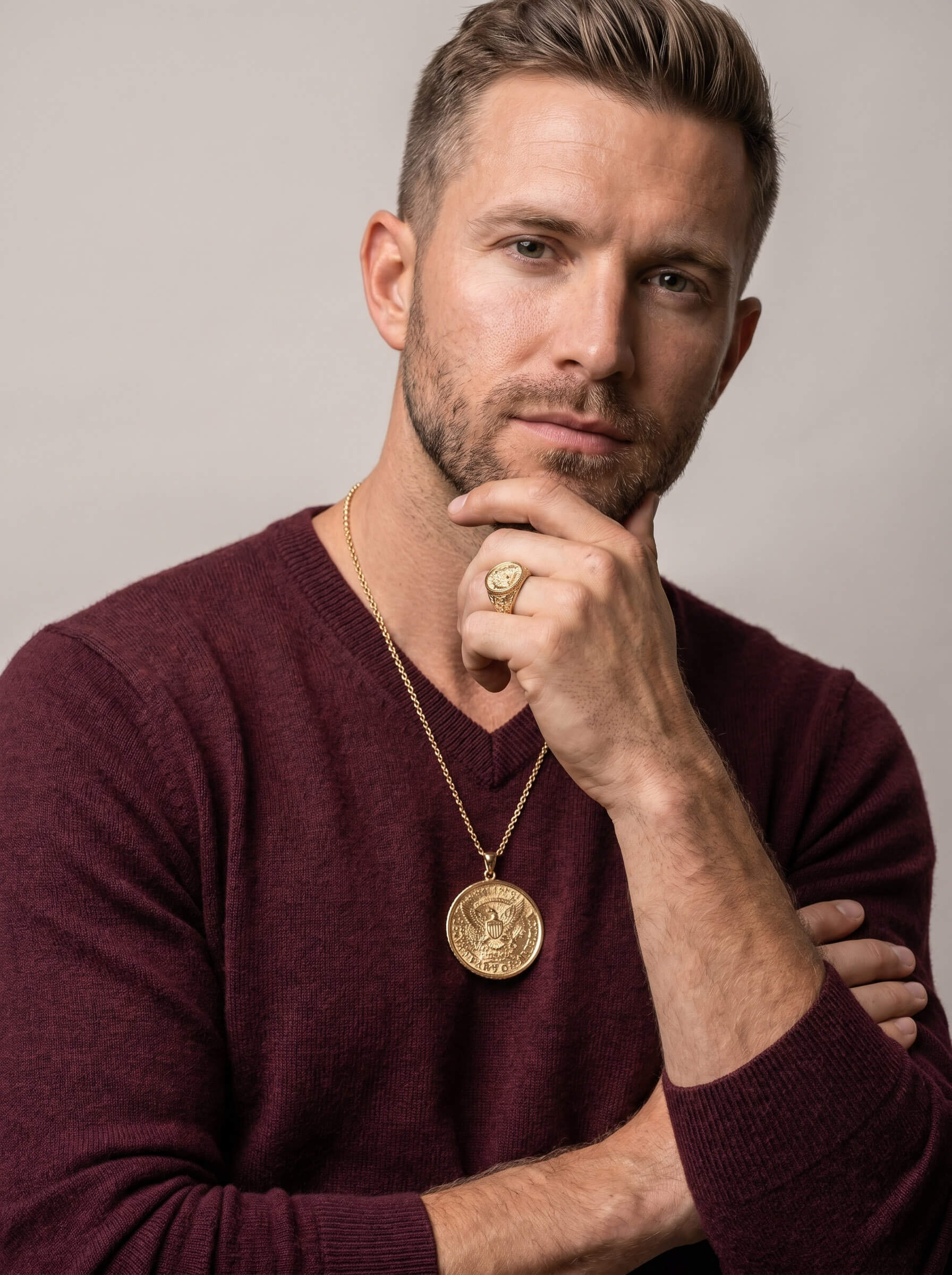 Man wearing a gold American Glory Pendant necklace and matching gold ring, dressed in a burgundy sweater, posing thoughtfully against a neutral background.
