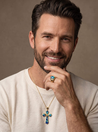 Man wearing a gold Wings Of Freedom Pendant necklace with blue opal-like inlays, smiling and showing a matching ring, dressed in a white textured shirt.