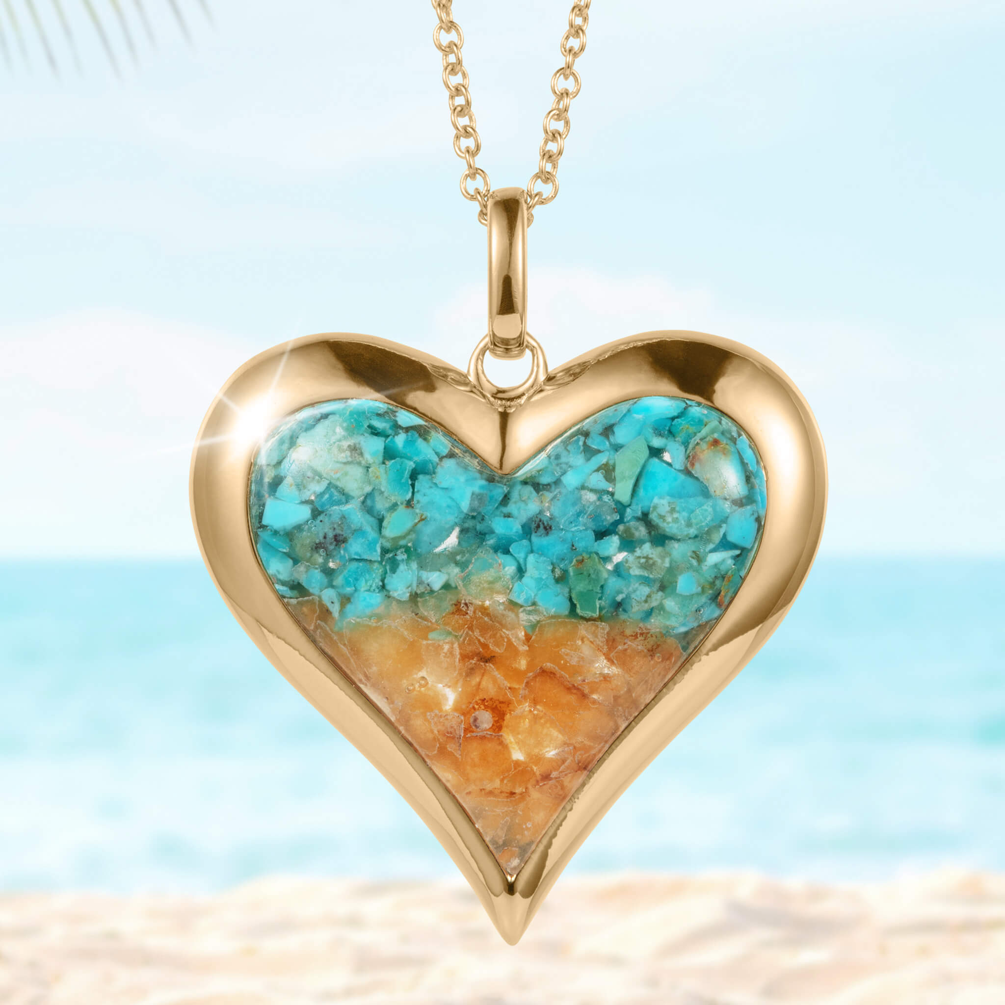 Heart-shaped gold pendant necklace from the Sunlit Shore Heart Collection featuring blue and sandy-colored stone inlay, displayed against a beach background.