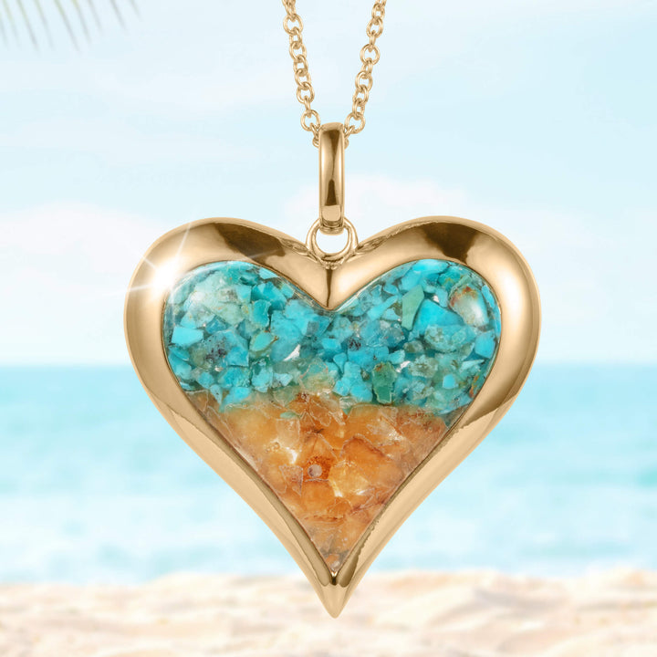 Heart-shaped gold pendant necklace from the Sunlit Shore Heart Collection featuring blue and sandy-colored stone inlay, displayed against a beach background.