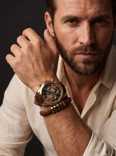Bearded man in a cream shirt wearing a brown leather chronograph watch and stacked bracelets while resting his hand near his face against a dark background.