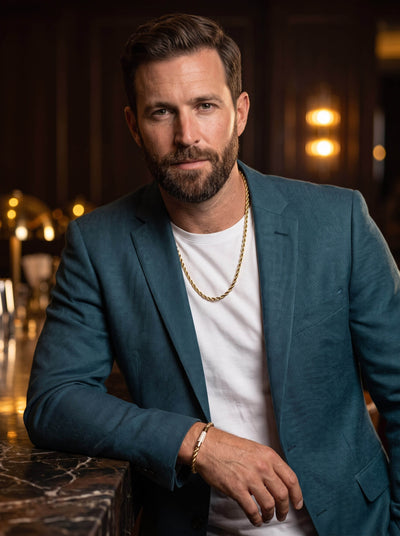 Bearded man in a teal blazer and white shirt wearing a gold chain necklace and bracelet while leaning against a marble bar in a warmly lit interior