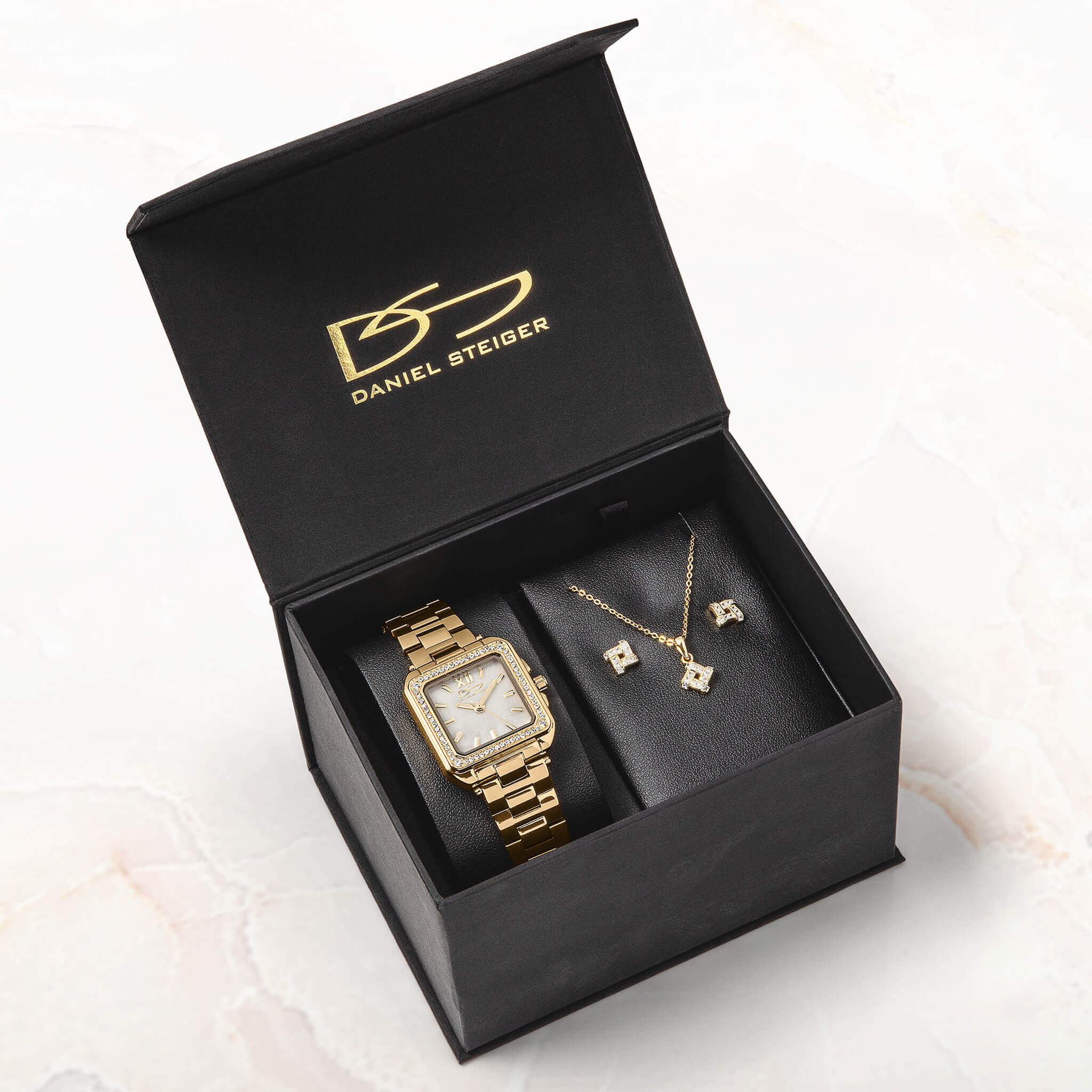 Open black Daniel Steiger presentation box displaying a gold-tone watch with a square face alongside a delicate necklace and matching earrings