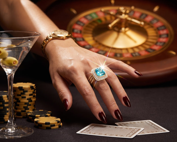 Ocean Flash Ladies Ring featuring a sparkling blue center stone in a gold tone setting worn on a hand beside casino chips cards and roulette wheel